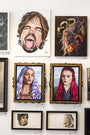 The display showcases diverse portrait artworks, including an oil on board by Gene Guynn. Top left is a caricature with an open mouth, followed by a fantasy dragon painting. Below are womens portraits with varied hair colors, surrounded by smaller sketches completing this art show tribute.