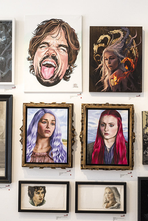 The display showcases diverse portrait artworks, including an oil on board by Gene Guynn. Top left is a caricature with an open mouth, followed by a fantasy dragon painting. Below are womens portraits with varied hair colors, surrounded by smaller sketches completing this art show tribute.