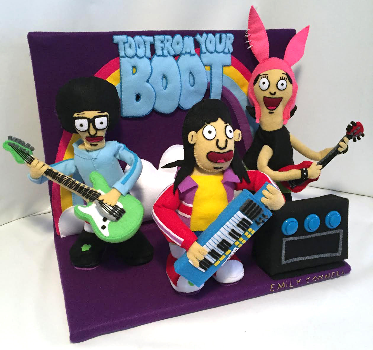 Felt figures of cartoon characters with instruments against a purple and rainbow backdrop, styled by Emily Connell. The text reads Toot From Your Boot, echoing the whimsical vibe similar to Spoke Art NYCs Bobs Burgers art show, creating a playful musical atmosphere.
