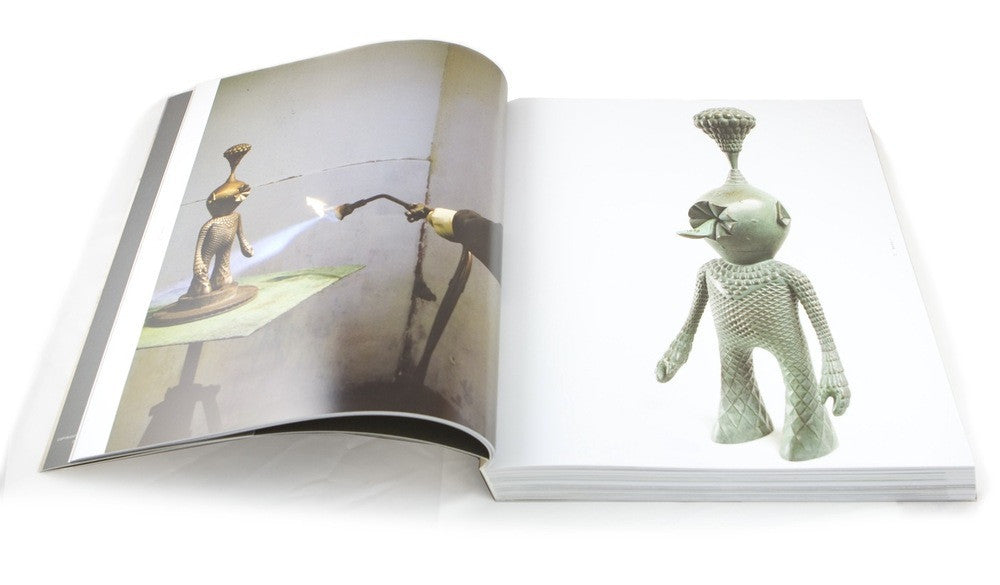 An open book reveals Mars-1s metallic humanoid sculpture: the left page shows welding or torching in action, while the right showcases the finished piece with its textured surface and distinctive headpiece.