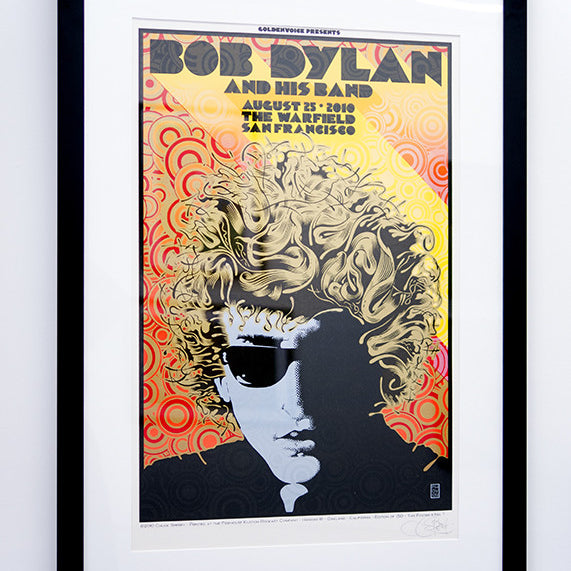 This custom framed print by Chuck Sperry showcases a psychedelic Bob Dylan concert art, featuring a stylized man with curly hair and sunglasses against a vibrant background. It includes concert details from August 25, 2010, in San Francisco.
