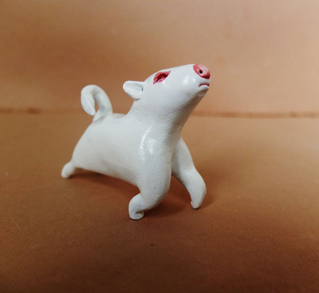 A small glossy white ceramic pig sculpture by Doubleparlour, featuring red eyes and a curly tail, stands on a light brown surface looking upwards.