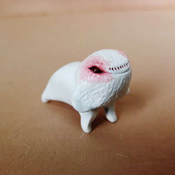 This hand-sculpted resin figure by Doubleparlour is a small, white creature with a textured surface, standing on four legs. It has a rounded body with a face featuring a pink patch and one dark eye against a light brown backdrop.