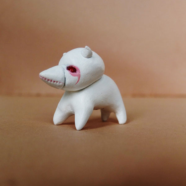 A small white figurine by Doubleparlour, crafted from resin with four legs, a pointed snout, two tiny horns, and red acrylic paint markings around its eyes. It sits on a plain light brown surface.