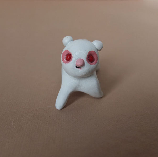 This small resin figurine from Doubleparlour, with large red eyes and a pink nose, resembles a panda and sits gracefully on a light brown surface, showcasing the brands distinctive artistry.