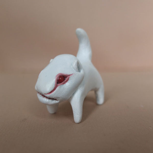 A small, white clay dinosaur figurine by Doubleparlour features red eyes and a long tail on a beige surface. The smooth, simplistic design stands out against the plain soft brown background, highlighting its hand-sculpted resin and acrylic paint details beautifully.