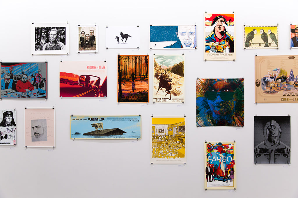 A gallery wall displays Joel Phillips posters and artworks, featuring film-themed pieces with diverse styles. Custom framing and UV-resistant plexiglass preserve the mix of portraits, abstract designs, and scenes on a pristine white wall.