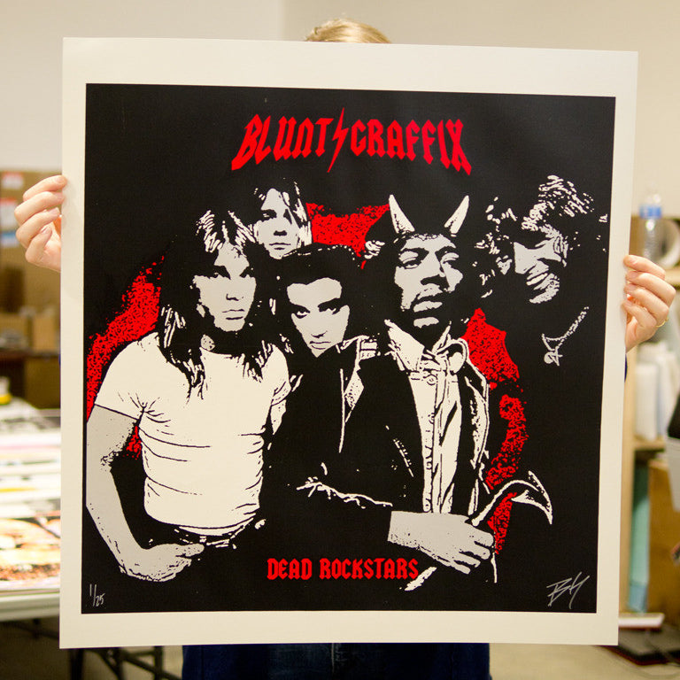 A person holds a striking screen print poster by Matt Dye, featuring illustrations of five musicians in a bold red and black color scheme, with BLUNT GRAFFIX at the top and DEAD ROCKSTARS at the bottom.