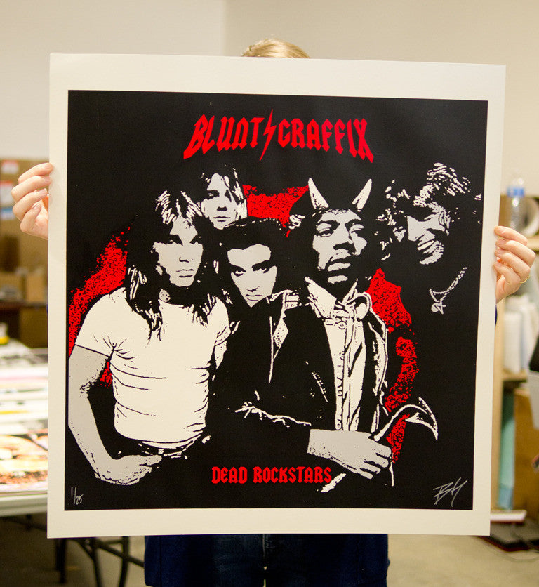 A person holds a striking screen print poster by Matt Dye, featuring illustrations of five musicians in a bold red and black color scheme, with BLUNT GRAFFIX at the top and DEAD ROCKSTARS at the bottom.