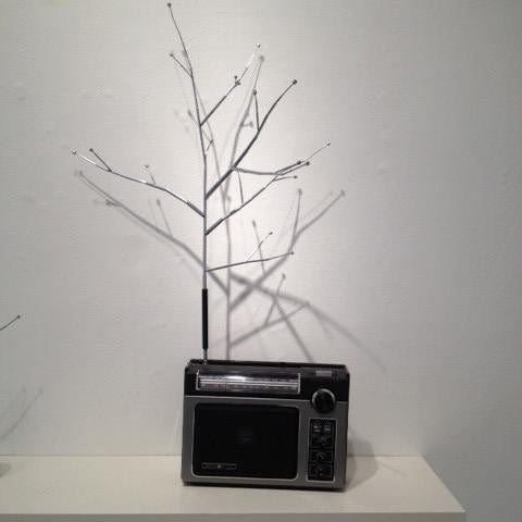 A small gray radio by Jeremiah Jenkins sits on a white shelf against a plain wall. A thin, bare branch with nodes extends from the top, casting intricate shadows and seamlessly blending mixed media elements for an evocative tableau.
