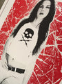 A woman with long hair sports a sleeveless Matt Dye top with a skull and lightning bolt design, echoing a Death Proof screenprint, and pairs it with shorts and a belt against a bold red and white abstract backdrop.