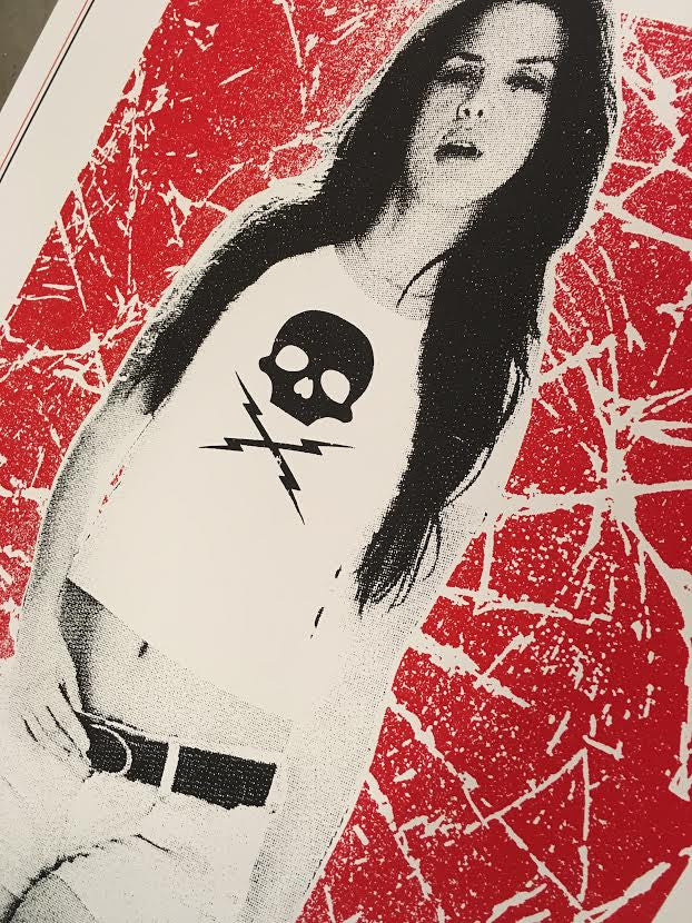 A woman with long hair sports a sleeveless Matt Dye top with a skull and lightning bolt design, echoing a Death Proof screenprint, and pairs it with shorts and a belt against a bold red and white abstract backdrop.