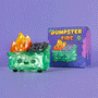 A translucent green Gem Dumpster Fire vinyl figure with orange crystal flames and a smiling face by 100% Soft sits before its colorful box on a vibrant purple background.