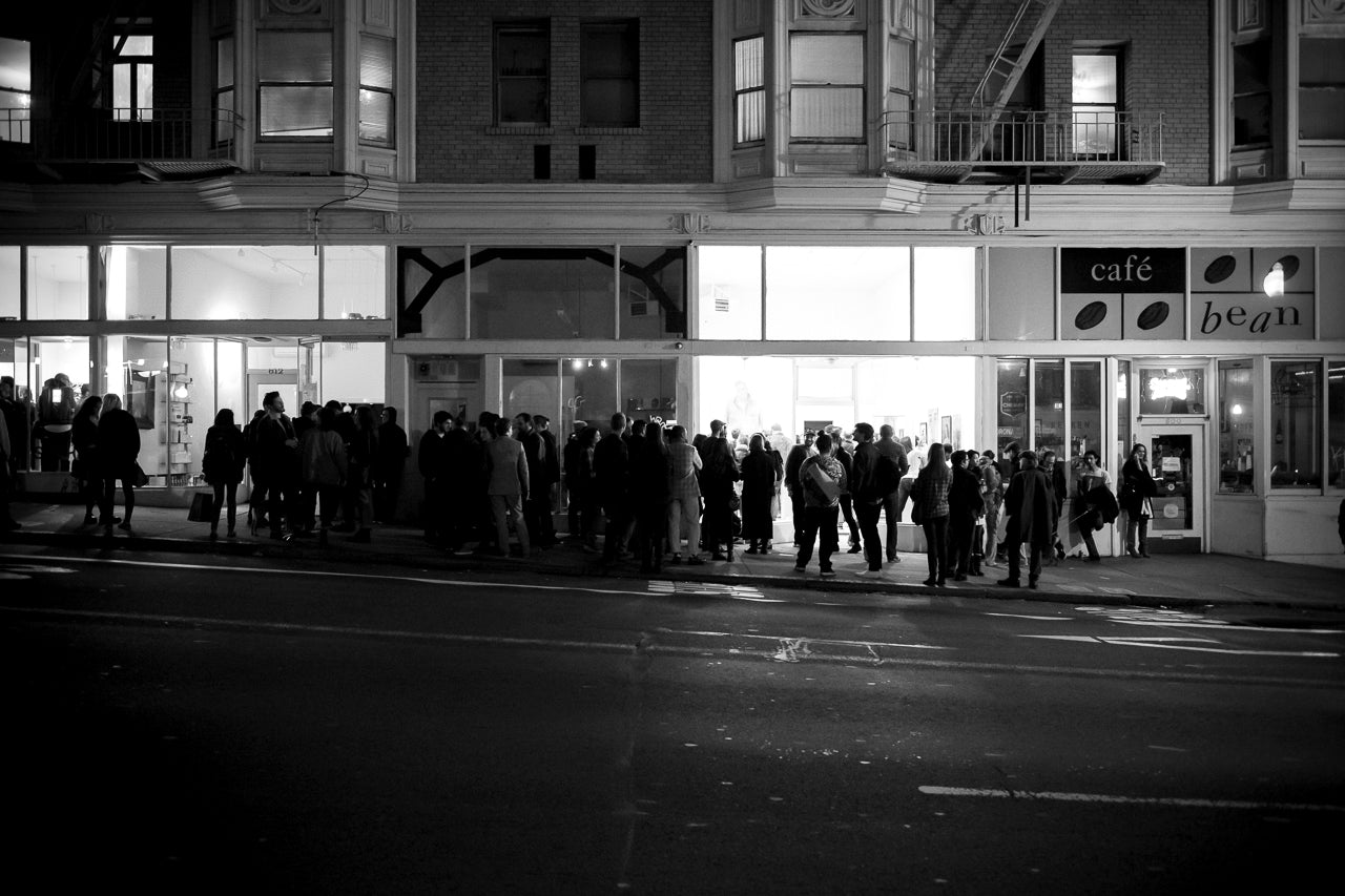 A black-and-white photo shows a crowd of people gathered outside Café Bean at night. The café windows are brightly lit, contrasting with the dark street. The building above has multiple windows and fire escapes.