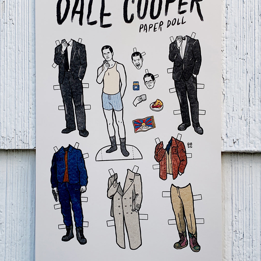 Brighton Ballard illustrated Dale Cooper paper doll sheet featuring a doll in underwear and various outfits, accessories, and faces to mix and match, all set against a white wood background—an original design.