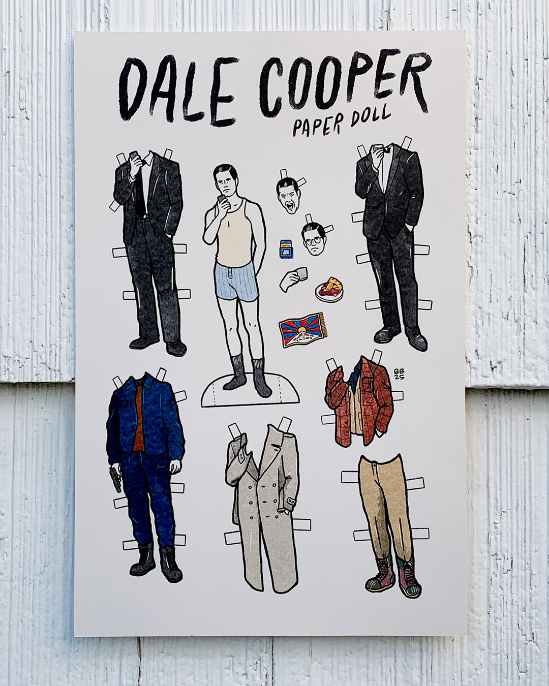 Brighton Ballard illustrated Dale Cooper paper doll sheet featuring a doll in underwear and various outfits, accessories, and faces to mix and match, all set against a white wood background—an original design.
