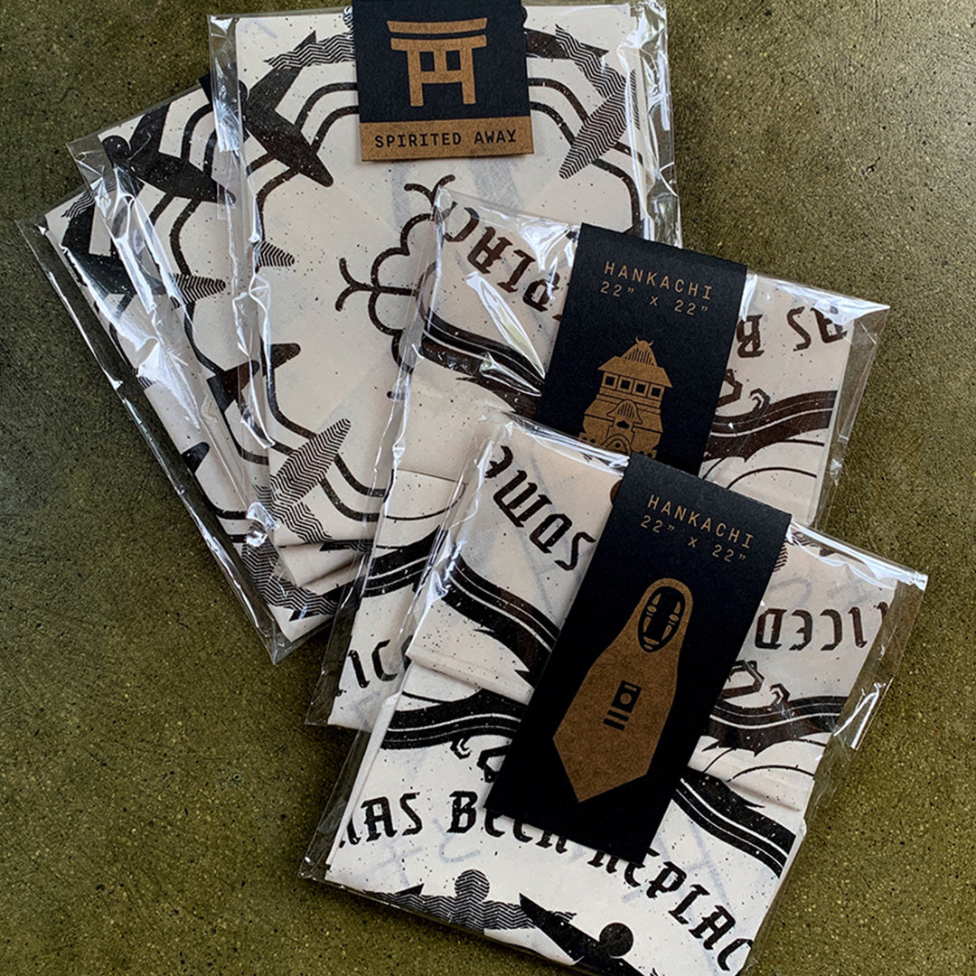 Three packages of folded white Derek Ballard handkerchiefs with black graphic designs rest on a brown surface. Each screen printed bandana includes a black label, some featuring a torii gate and Spirited Away references.