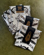 Three packages of folded white Derek Ballard handkerchiefs with black graphic designs rest on a brown surface. Each screen printed bandana includes a black label, some featuring a torii gate and Spirited Away references.