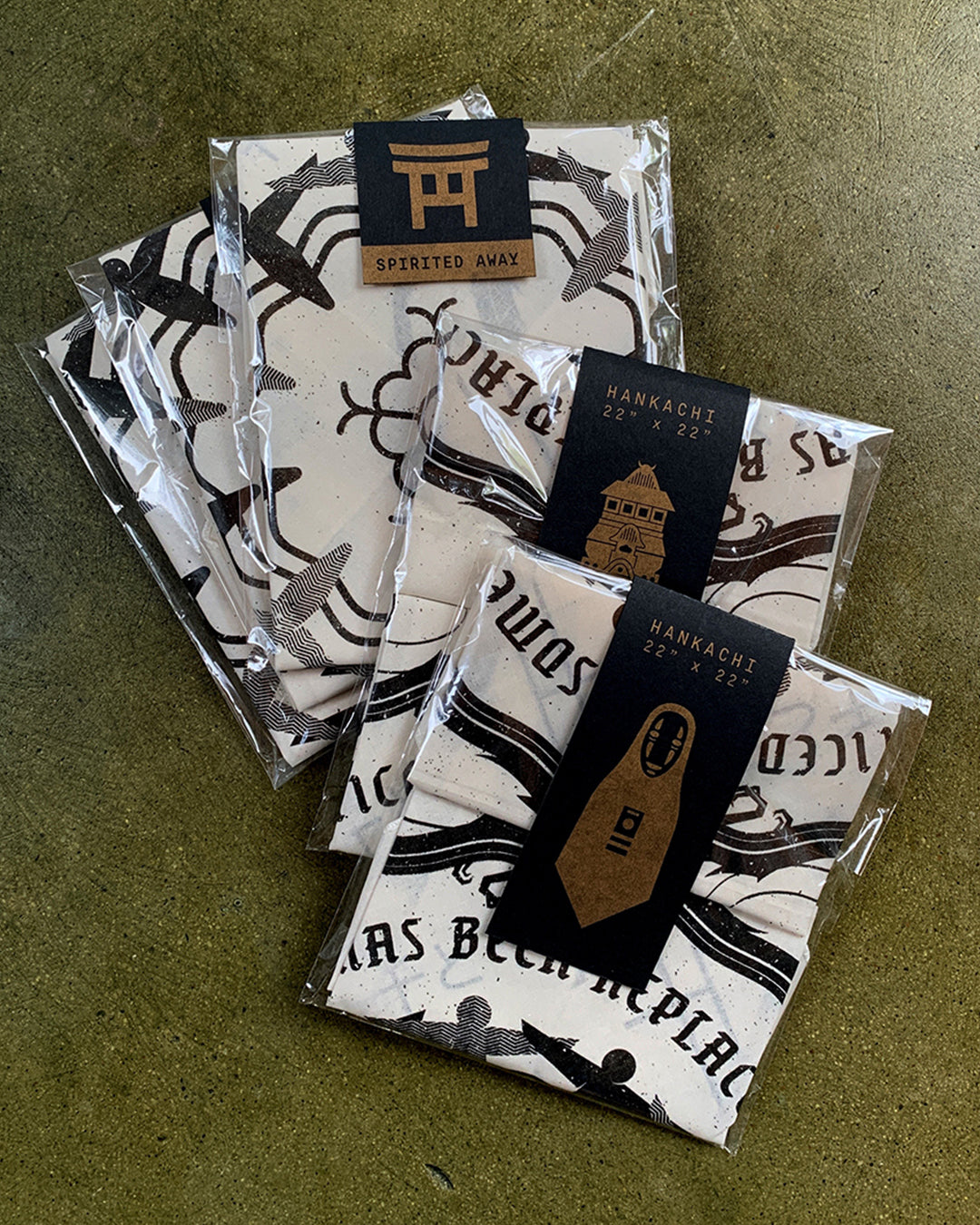 Three packages of folded white Derek Ballard handkerchiefs with black graphic designs rest on a brown surface. Each screen printed bandana includes a black label, some featuring a torii gate and Spirited Away references.