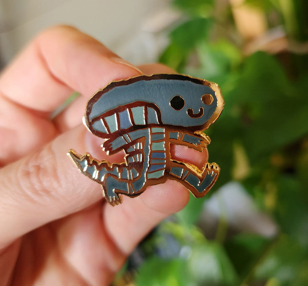 A hand holds a cute, cartoon-style Scott C. hard enamel pin featuring a smiling blue-grey trilobite with arms and legs, with blurred green plants in the background.