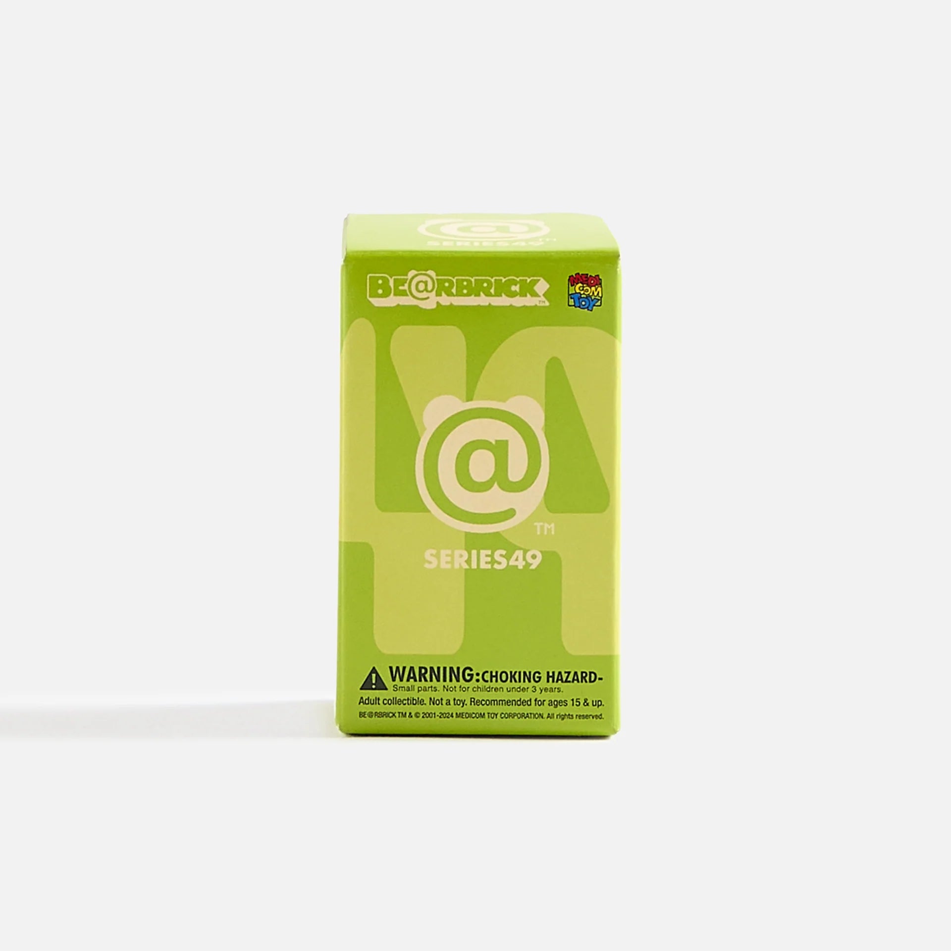 A green Medicom Be@rbrick Series 49 blind box stands upright against a plain white background. The box features the @ symbol, the series number, and a warning label about choking hazards.