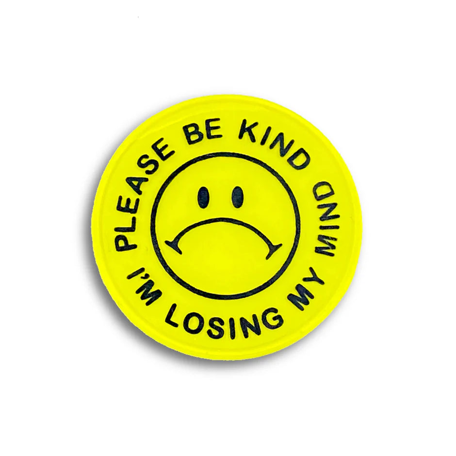 A yellow circular hard enamel pin by Spoke Art features a black sad face and “PLEASE BE KIND I’M LOSING MY MIND” around the edge—a limited edition badge, perfect for fans of Special Ed Toys.