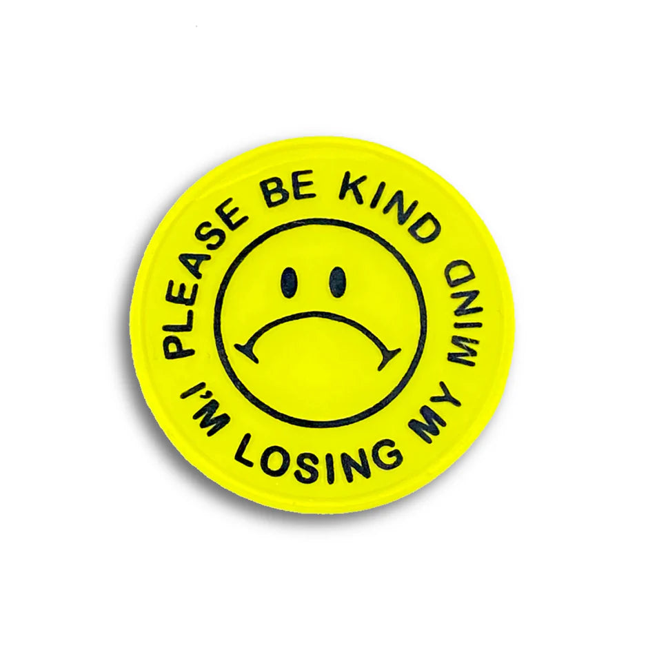 A yellow circular hard enamel pin by Spoke Art features a black sad face and “PLEASE BE KIND I’M LOSING MY MIND” around the edge—a limited edition badge, perfect for fans of Special Ed Toys.
