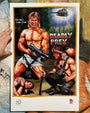 A hand holds a Deadly Prey digital print, showing a vintage-style movie poster with a muscular man in torn shorts holding a gun, a defeated man on the ground, another armed figure, and a helicopter above an explosion.