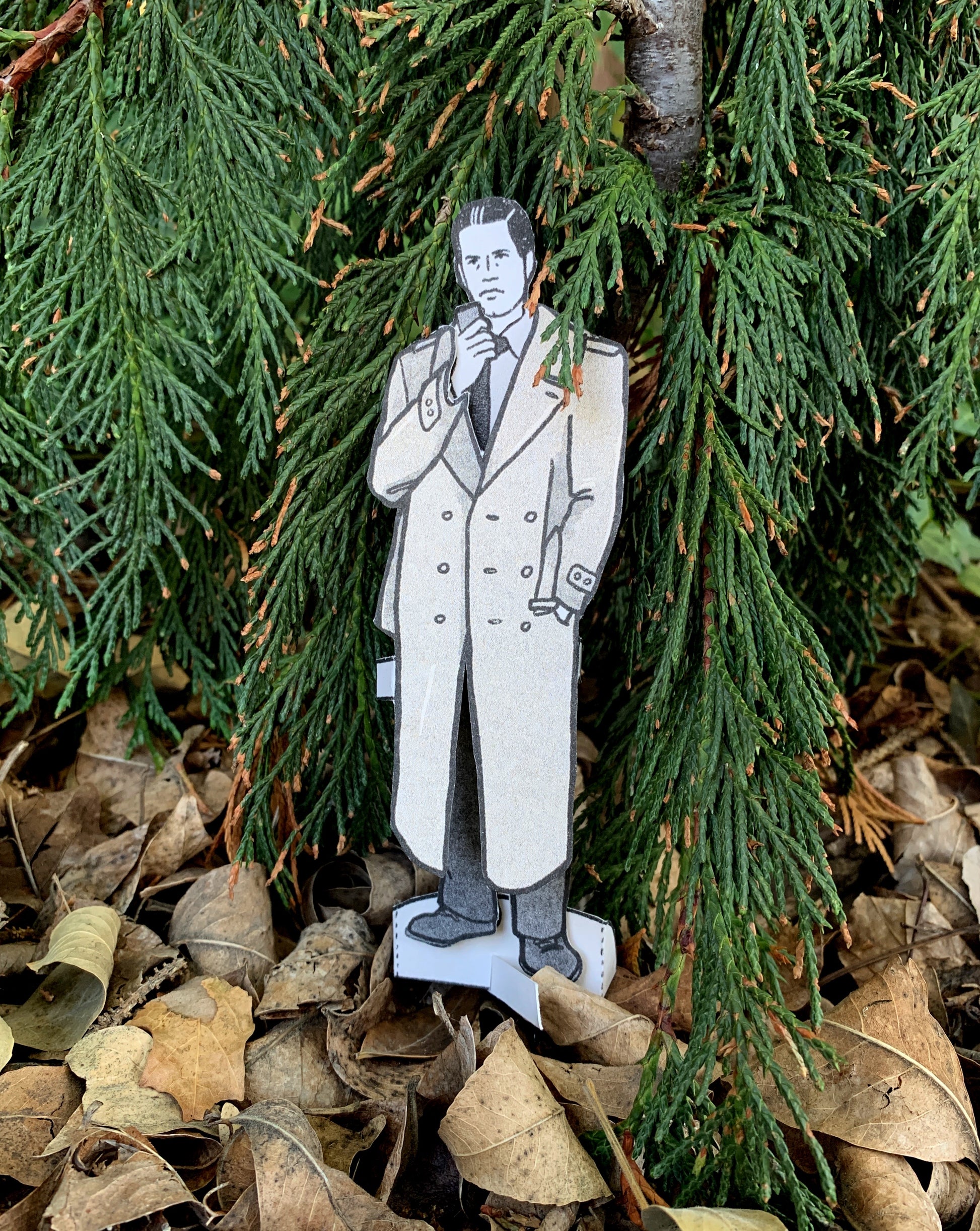 Cardboard cutout of a man in a coat standing among green foliage and fallen leaves.