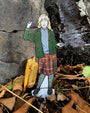 Paper figure of a person in a green jacket and plaid skirt standing among rocks and leaves.
