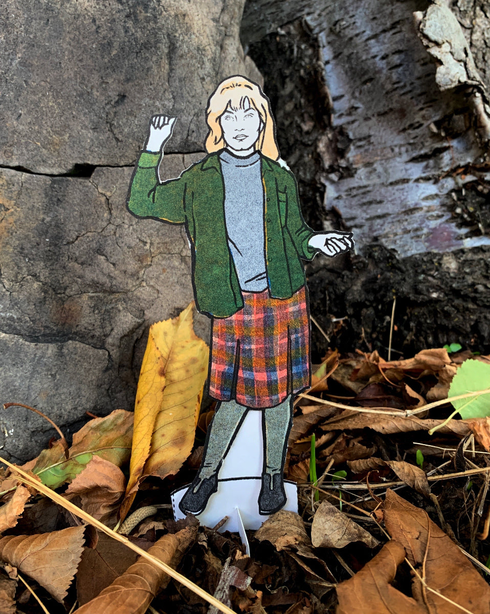 Paper figure of a person in a green jacket and plaid skirt standing among rocks and leaves.