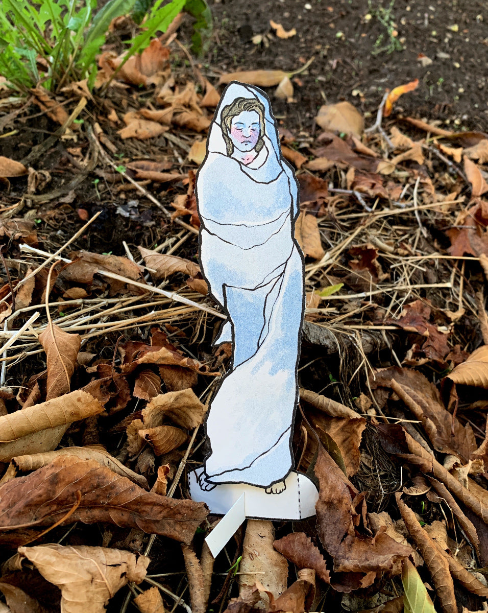 Cardboard cutout of a figure wrapped in white cloth on dry leaves