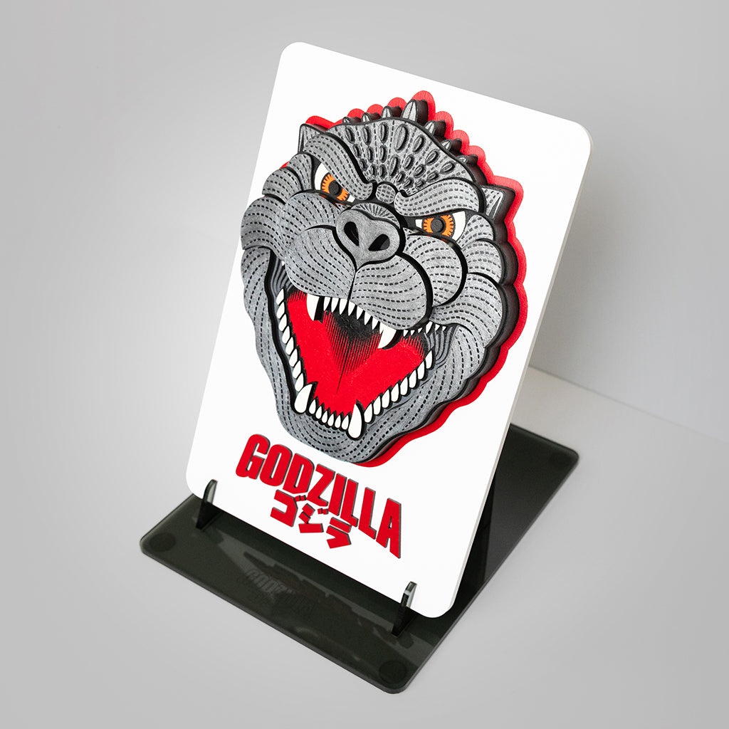 Mixed media sculpture of an enlarged enamel pin of Godzilla's face on a white backing with the Godzilla logo in red underneath the pin on a plastic stand