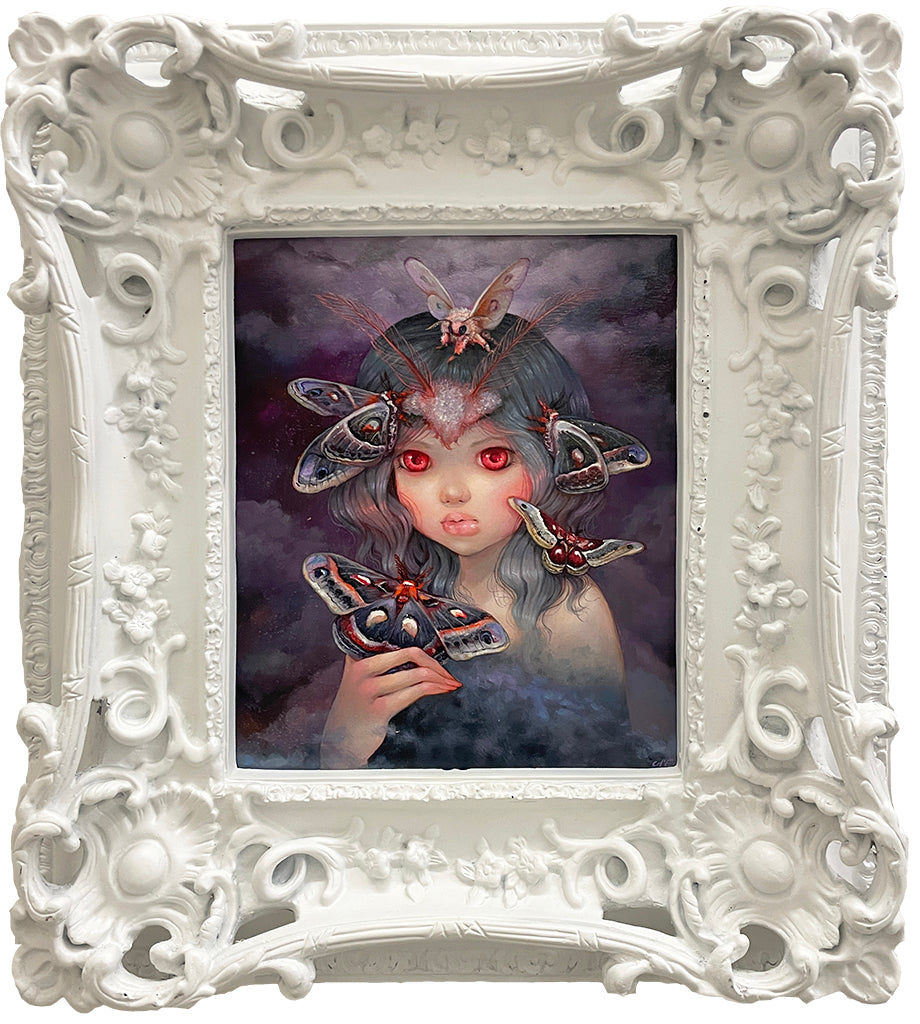 A Spoke Art framed painting in Camilla d'Errico's style shows a girl with large red eyes and dark, wavy hair among colorful moths. The ornate white floral frame enhances the mysterious, surreal vibe of Prophet Of The Night.