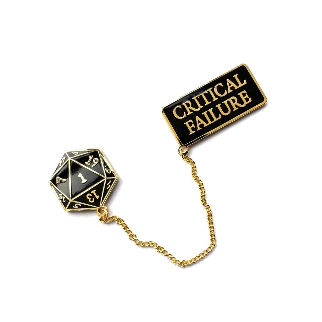 Spoke Art’s limited edition hard enamel pin set in black and gold features a 20-sided die showing 1, linked by a chain to a rectangular gold-plated pin reading “CRITICAL FAILURE” in shimmering gold letters.
