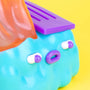 A close-up of a colorful limited edition 100% Soft vinyl figure featuring a blue bumpy surface, purple and white oval eyes and mouth, a translucent orange fin, and a purple panel, set against a bright yellow background.