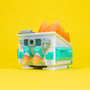 A 100% Soft Mecha Dumpster Fire vinyl figure, designed like a futuristic building with teal accents and orange rocket boosters, sits on a bright yellow background with flames erupting from its roof—a unique collectible debuting at San Diego Comic-Con 2023.