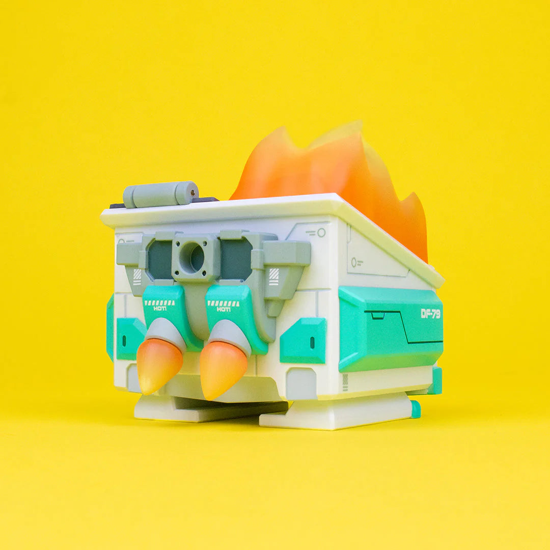 A 100% Soft Mecha Dumpster Fire vinyl figure, designed like a futuristic building with teal accents and orange rocket boosters, sits on a bright yellow background with flames erupting from its roof—a unique collectible debuting at San Diego Comic-Con 2023.