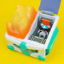 A toy mouse wearing a helmet sits inside a futuristic, teal and white Mecha Dumpster Fire vehicle with an orange translucent cover, all set against a bright yellow background—this must-have vinyl figure from 100% Soft is inspired by San Diego Comic-Con 2023.