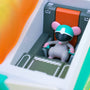 A small vinyl figure by 100% Soft depicts a toy mouse wearing a teal helmet and black visor seated in a futuristic vehicle cockpit with orange and gray controls, inspired by the Mecha Dumpster Fire revealed at San Diego Comic-Con 2023.