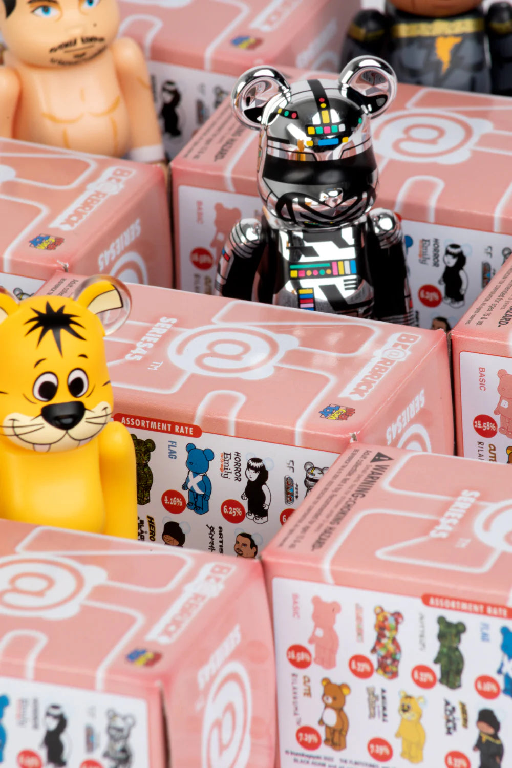 Colorful bear and animal figurines are displayed among pink Blind Box packages featuring images and rarity rates for collectible toys. The focus is on a shiny robot bear from the Bearbrick Series 45 by Medicom and a yellow cartoon tiger character.