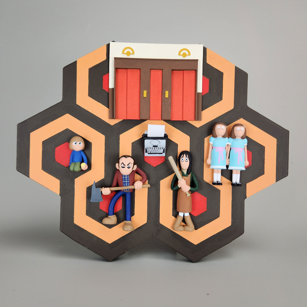 Spoke Art’s clay figures from The Shining stand on a geometric carpet base, featuring Jack with an axe, Wendy with a bat, Danny, the twins, elevator doors splashed red, and a typewriter—iconic elements from the film.
