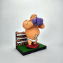 Geoff Trapp’s cartoon boxer figurine, with purple gloves, bandages, and white trunks, stands on a platform before a painted crowd—its worried face and rounded features evoke King Hippo from Mike Tyson’s Punchout.