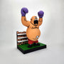 A cartoonish, bald boxer with purple gloves and red shorts celebrates victory on a green platform before a small crowd—an ideal Geoff Trapp piece for fans of video game-inspired art and King Hippo designs.