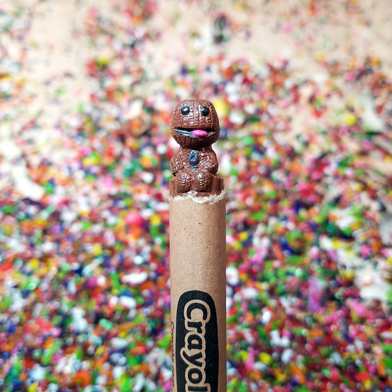A Hoang Tran brown crayon is sculpted at the tip into a tiny figure resembling a stitched doll, inspired by LittleBigPlanet art, standing against a blurred background of colorful crayon shavings.