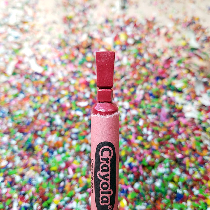 A red Hoang Tran crayon with a flawless squared tip stands upright in sharp focus, set against a blurred background of multicolored crayon shavings—a striking example of crayon art.