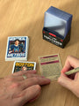 A person writes on the back of a Cuyler Smith trading card with a pen. Nearby are a stack of trading cards, a box of Ultra PRO Toploader card sleeves, and a few more cards from the Heated Rivalry set on a wooden table—perhaps preparing to create a signed numbered edition.