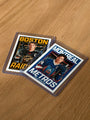 Two hockey trading cards from the Culyer Smith trading cards Heated Rivalry set, each in protective sleeves, are lying on a wooden surface. One card reads Boston Raiders and the other Montreal Metros, each showing a hockey player in uniform holding a hockey stick. This signed numbered edition perfectly captures the spirit of classic competition.