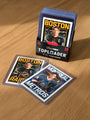 Three Culyer Smith trading cards from the Heated Rivalry set are displayed in protective sleeves on a wooden surface, two laid out and one standing in a box. The cards feature illustrated players from Boston and Montreal teams, making this signed numbered edition a standout collector’s piece.