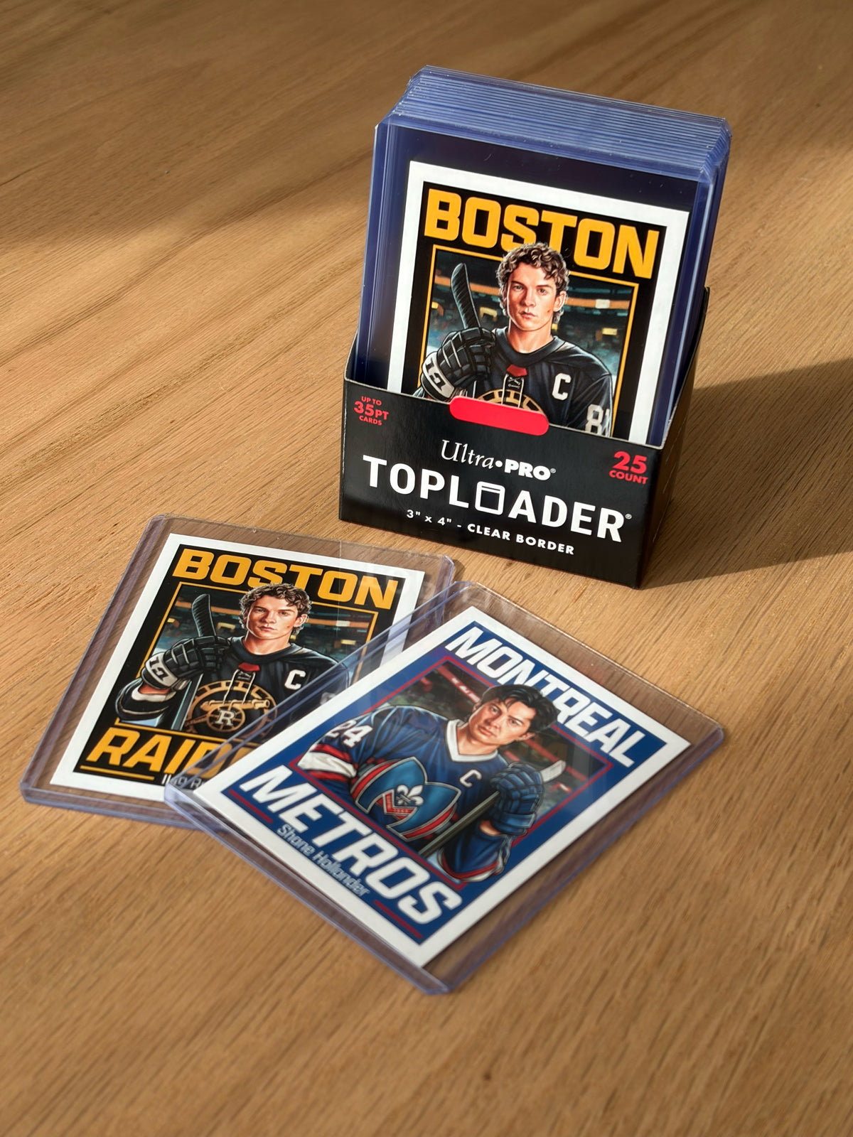 Three Culyer Smith trading cards from the Heated Rivalry set are displayed in protective sleeves on a wooden surface, two laid out and one standing in a box. The cards feature illustrated players from Boston and Montreal teams, making this signed numbered edition a standout collector’s piece.